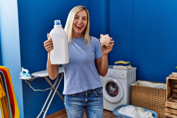 Beautiful woman doing laundry holding detergent bottle and piggy bank smiling and laughing hard out loud because funny crazy joke.  - Foto, Bild
