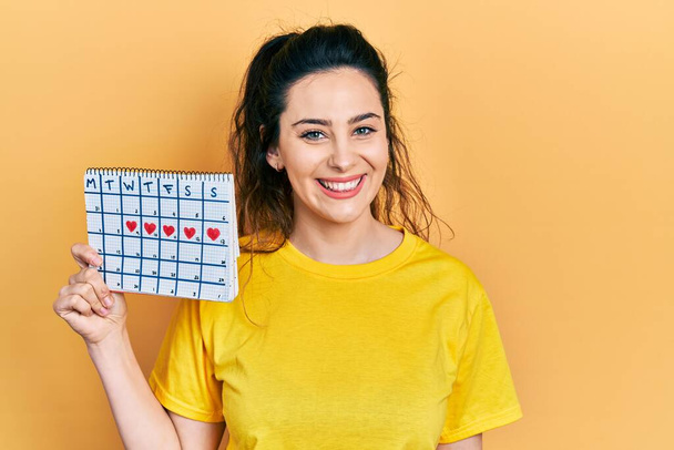 Jeune femme hispanique tenant calendrier cardiaque regardant positif et heureux debout et souriant avec un sourire confiant montrant les dents  - Photo, image