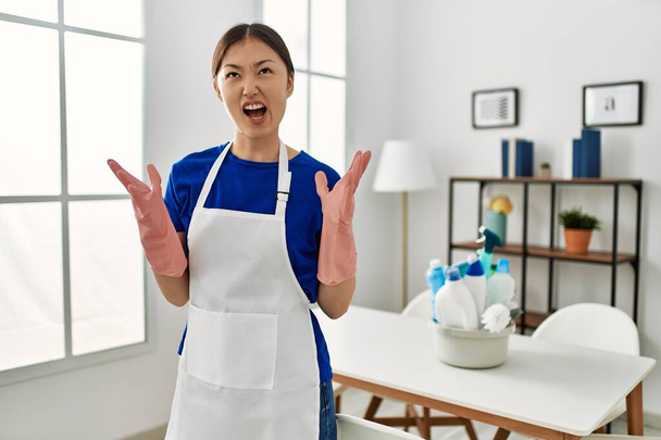 Young chinese girl wearing cleaner uniform standing at home crazy and mad shouting and yelling with aggressive expression and arms raised. frustration concept.  - Fotó, kép