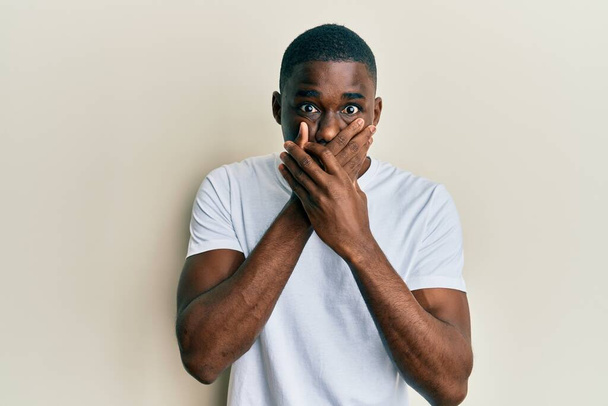 Ein junger afrikanisch-amerikanischer Mann in einem lässigen weißen T-Shirt schockierte, als er fälschlicherweise den Mund mit den Händen verdeckte. Geheimes Konzept.  - Foto, Bild