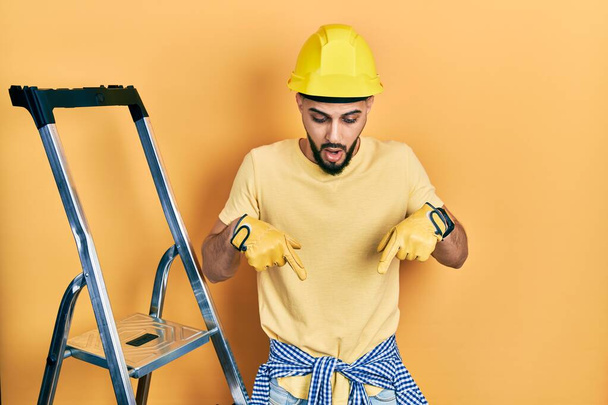 Handsome man with beard by construction stairs wearing hardhat pointing down with fingers showing advertisement, surprised face and open mouth  - Photo, Image