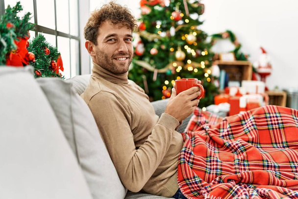 Young hispanic man drinking coffee sitting by christmas tree at home - Foto, afbeelding