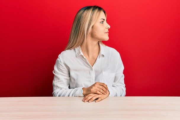 Jeune femme caucasienne portant des vêtements décontractés assis sur la table regardant vers le côté, pose de profil relax avec visage naturel avec sourire confiant.  - Photo, image