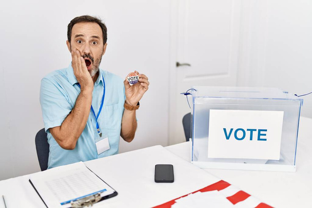 Middle age man with beard sitting by ballot holding i vote badge afraid and shocked, surprise and amazed expression with hands on face  - Foto, afbeelding