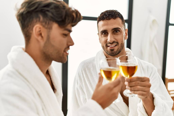 Two hispanic men couple toasting with champagne sitting on massage table at beauty center - Foto, afbeelding