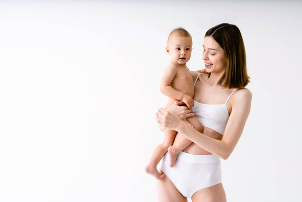 Happy woman holding her baby  on colored background -  Young woman wearing underwear taking care of her little son  - Pregnancy, motherhood, people and expectation concept - Photo, image
