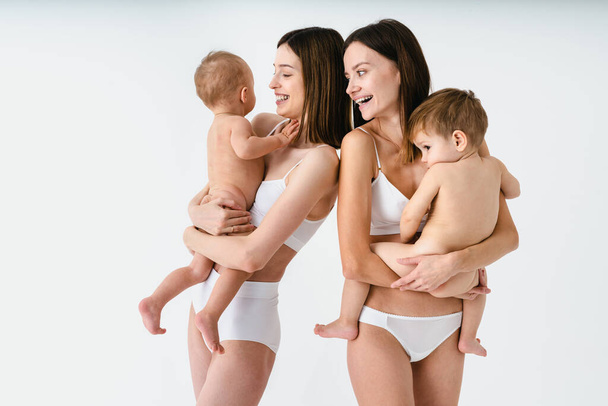 Mujeres felices sosteniendo a sus bebés sobre un fondo de color - Mujeres jóvenes usando ropa interior cuidando a sus hijos pequeños - Embarazo, maternidad, personas y concepto de expectativa - Foto, imagen