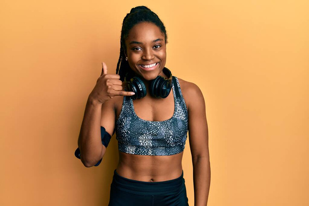 African american woman with braided hair wearing sportswear and arm band smiling doing phone gesture with hand and fingers like talking on the telephone. communicating concepts.  - Foto, immagini