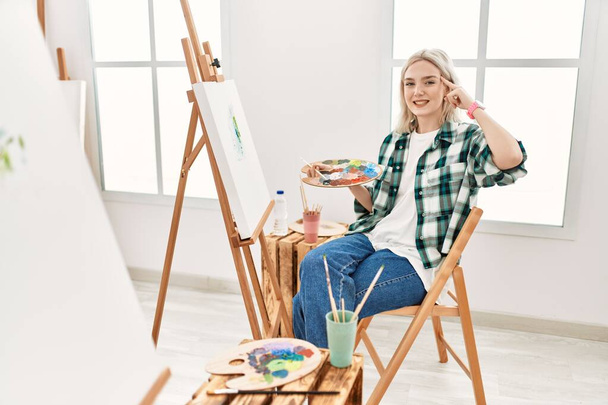 Молода художниця, яка малює на полотні в художній студії, посміхається, вказуючи на голову одним пальцем, чудовою ідеєю чи думкою, хорошою пам'яттю
  - Фото, зображення