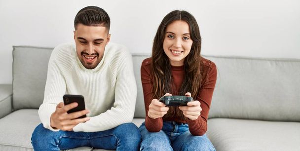 Νεαρό ζευγάρι ισπανόφωνων παίζει βιντεοπαιχνίδι και χρησιμοποιεί smartphone στο σπίτι. - Φωτογραφία, εικόνα