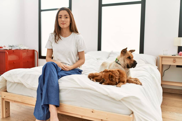 Young brunette woman sitting on the bed with two dogs with serious expression on face. simple and natural looking at the camera.  - Foto, Imagem