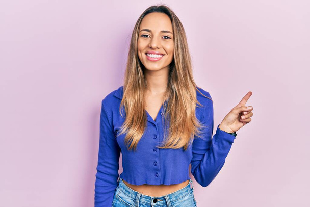 Hermosa mujer hispana vistiendo una camisa azul casual con una gran sonrisa en la cara, señalando con el dedo de la mano hacia un lado mirando a la cámara.  - Foto, Imagen