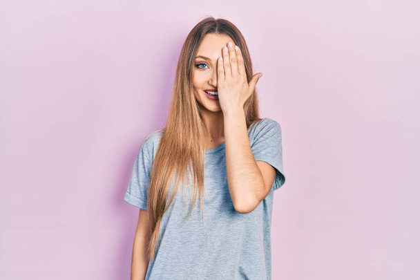 Young blonde girl wearing casual t shirt covering one eye with hand, confident smile on face and surprise emotion.  - Fotografie, Obrázek