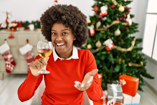 jong afrikaans amerikaans vrouw toasten met wijn zitten door kerstboom thuis - Foto, afbeelding