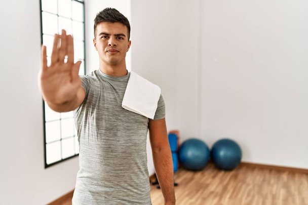 Young hispanic man wearing sportswear and towel at the gym doing stop sing with palm of the hand. warning expression with negative and serious gesture on the face.  - Фото, изображение