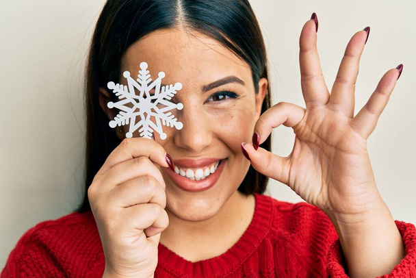 Beautiful brunette woman holding snowflake over eye doing ok sign with fingers, smiling friendly gesturing excellent symbol  - Fotoğraf, Görsel