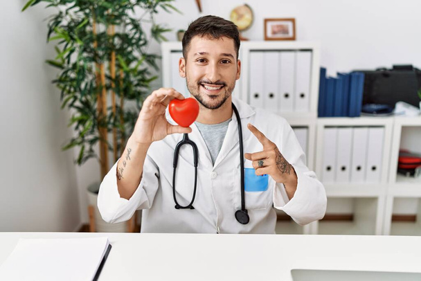 Young doctor holding heart at medical clinic smiling happy pointing with hand and finger  - Foto, immagini