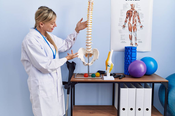 Jonge blonde vrouw draagt fysiotherpist uniform wijzend naar anatomisch model van wervelkolom bij fysiotherapie kliniek - Foto, afbeelding