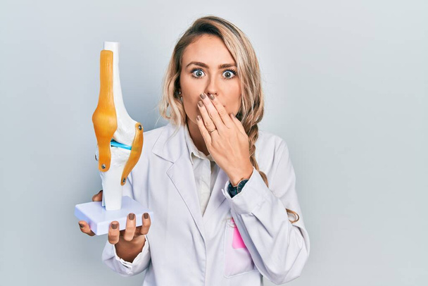 Beautiful young blonde doctor woman holding anatomical model of knee joint covering mouth with hand, shocked and afraid for mistake. surprised expression  - Valokuva, kuva
