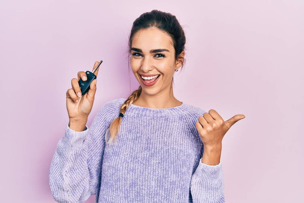 Young hispanic girl holding key of new car pointing thumb up to the side smiling happy with open mouth  - 写真・画像