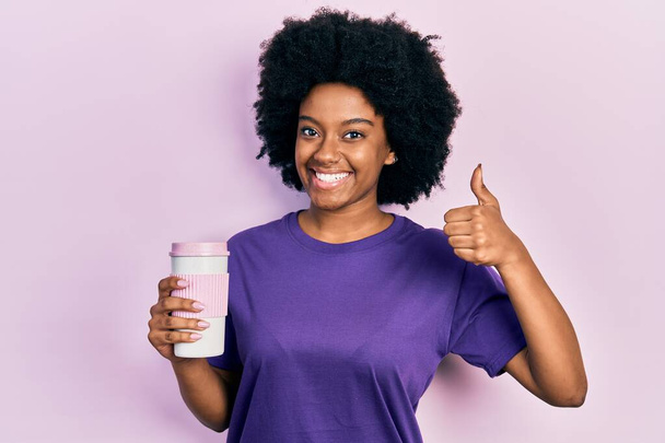 Νεαρή Αφροαμερικανή γυναίκα που κρατάει τον καφέ χαμογελώντας χαρούμενη και θετική, με τον αντίχειρα να κάνει εξαιρετική δουλειά και το σήμα έγκρισης  - Φωτογραφία, εικόνα