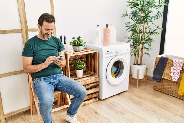 Middle age hispanic man doing laundry using smartphone waiting at home. - Foto, Bild