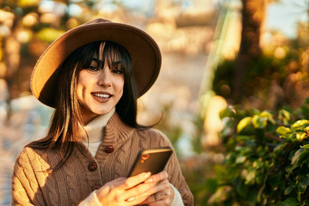 Brunette femme portant un chapeau d'hiver souriant en utilisant un smartphone au parc - Photo, image