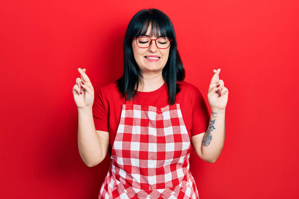 Young hispanic woman wearing cook apron and glasses gesturing finger crossed smiling with hope and eyes closed. luck and superstitious concept.  - Foto, imagen