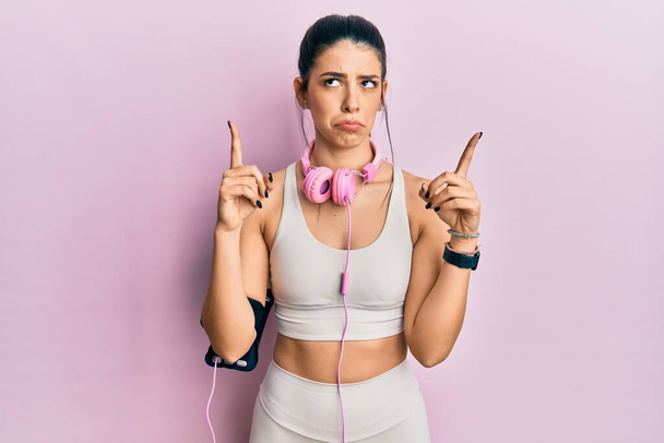 Young hispanic woman wearing gym clothes and using headphones pointing up looking sad and upset, indicating direction with fingers, unhappy and depressed.  - Fotó, kép