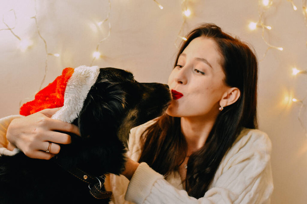 A shot of a Caucasian girl kissing her dog with Santa hat and Christmas lights on the wall - Foto, afbeelding