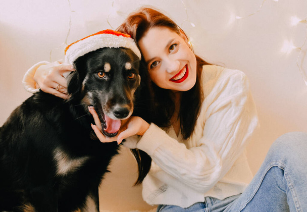 A closeup shot of a Caucasian girl smiling to the camera with her dog and Christmas lights on wall - Foto, Imagen