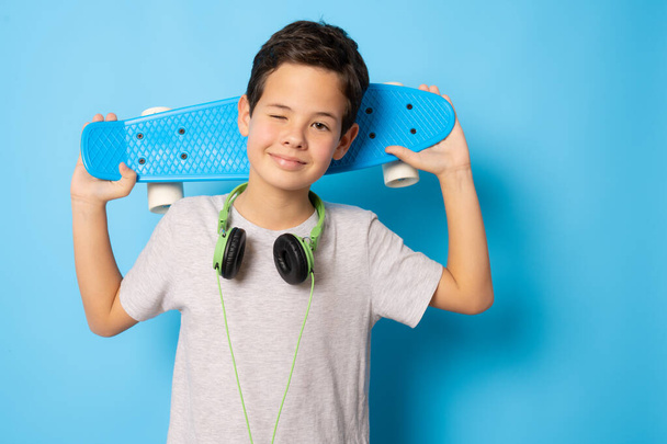 Хлопчик у повсякденному одязі, що тримає скейтборд, стоїть ізольовано на синьому фоні
. - Фото, зображення