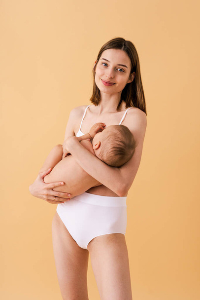 Femme heureuse tenant son bébé sur fond coloré - Jeune femme portant des sous-vêtements prenant soin de son petit fils - Grossesse, maternité, personnes et concept d'attente - Photo, image