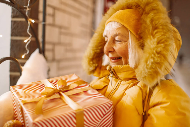 Compras de vacaciones. Foto de alegre mujer madura de los años 50 usando ropa de abrigo sosteniendo la caja de regalo de Navidad para un evento especial en una ciudad en invierno.. - Foto, Imagen