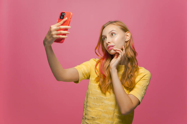 Pretty young girl with perfect glowing healthy skin posing doing selfie on phone on pink studio background - Foto, imagen