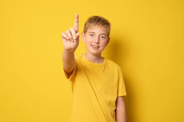 Usmívající se chlapec v ležérní tričko ukazující prstem nahoru stojící izolovaně přes žluté pozadí. - Fotografie, Obrázek