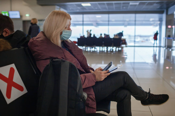医療用マスクのソロ旅行者は、空港でのフライトを待っている間、ターミナルでモバイルを使用します。 - 写真・画像