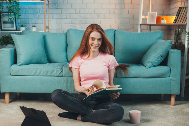 Caucasian woman studying at home, sitting on floor and reading diary. Red head woman. Smiling face. Quarantine lifestyle. Coronavirus - Fotografie, Obrázek