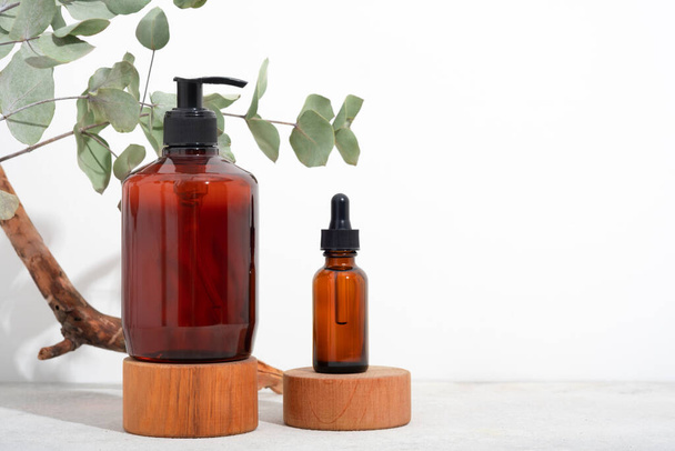 Mockup Spa body cosmetics in brown glass bottles serum with a pipette, oil, gel, shower soap, shampoo with a pump on a wooden pedestal and a white background with eucalyptus branches. Copy space - Photo, Image