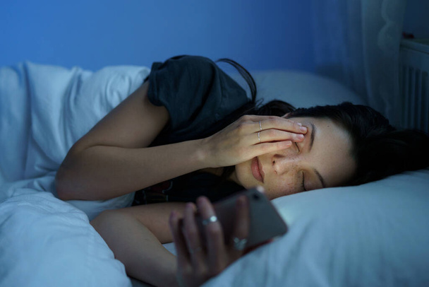 Yorgun bir kadın akıllı telefon ekranına bakıyor, gece geç saatte yatakta yatıyor. Sosyal medya bağımlılığı - Fotoğraf, Görsel
