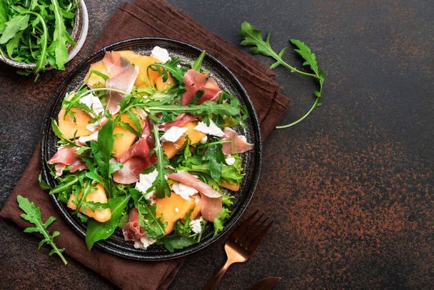 Summer salad with melon, jamon, white cheese and arugula on brown table background, top view, copy space - Foto, Bild
