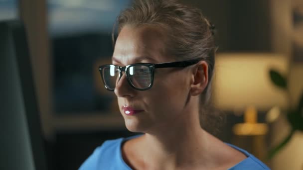 Woman in glasses looking on the monitor and surfing Internet. The monitor screen is reflected in the glasses. Work at night. Home Office. Remote work - Footage, Video