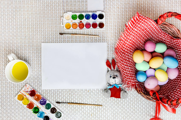 Painted Easter eggs and a toy bunny lie in a wooden basket on a wooden table by the window next to a white sheet with space for text. Top View Mocup. Selective focus and white background. - Photo, Image