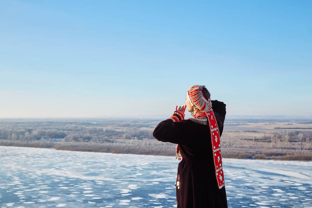 伝統的なドレスやヘッドギアで匿名の女性のバックビューは、顔に触れ、自然の寒い冬の日に青い空に対して凍結川を賞賛します - 写真・画像