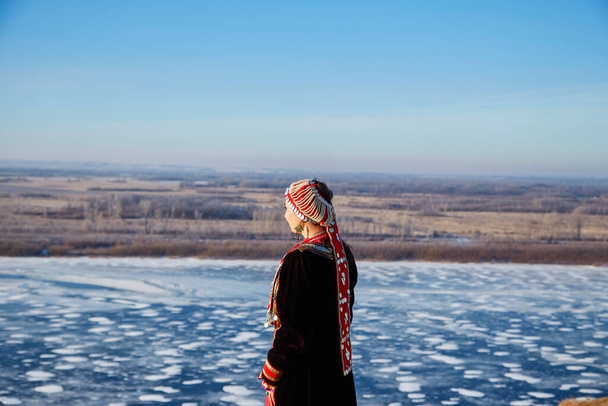 Back view of anonymous female in traditional dress and headgear touching face and admiring frozen river against blue sky on cold winter day in nature - Photo, Image