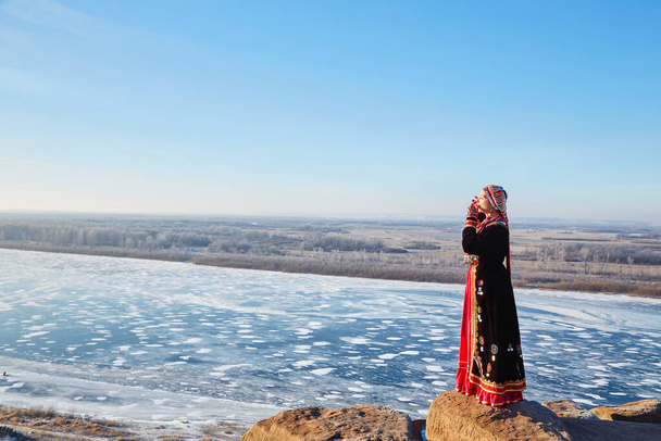 Side view of woman in authentic costume is playing a musical instrument jew's harp with the frozen river on the background while standing on stones near the village on a cloudless day in the countrysid - Photo, Image