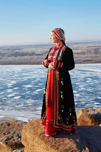 A young woman stands on the edge of a cliff in winter dressed in a national Bashkir costume - Foto, Imagem