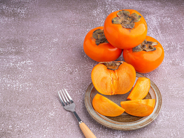 Fresh ripe persimmons with slices of persimmon on a plate with cutlery and whole fruit on gray stone background. Space for text. Close-up photo. Concept of health fruit. - Fotoğraf, Görsel