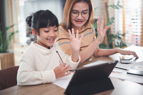 Asiatisches Mädchen, das ein Tablet benutzt, um mit ihrer Mutter Online-Technologie zu lernen und nebenbei zu beraten. Konzept der Online-Bildung, soziales Fernlernen zu Hause während Quarantäne und Schulferien. - Foto, Bild