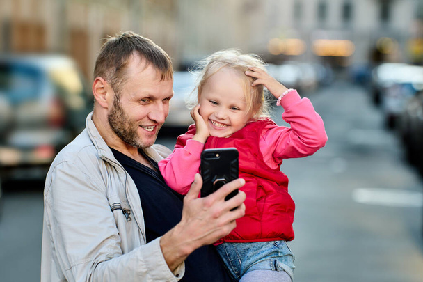 Father and daughter look at screen of handy outdoors and smile. - 写真・画像